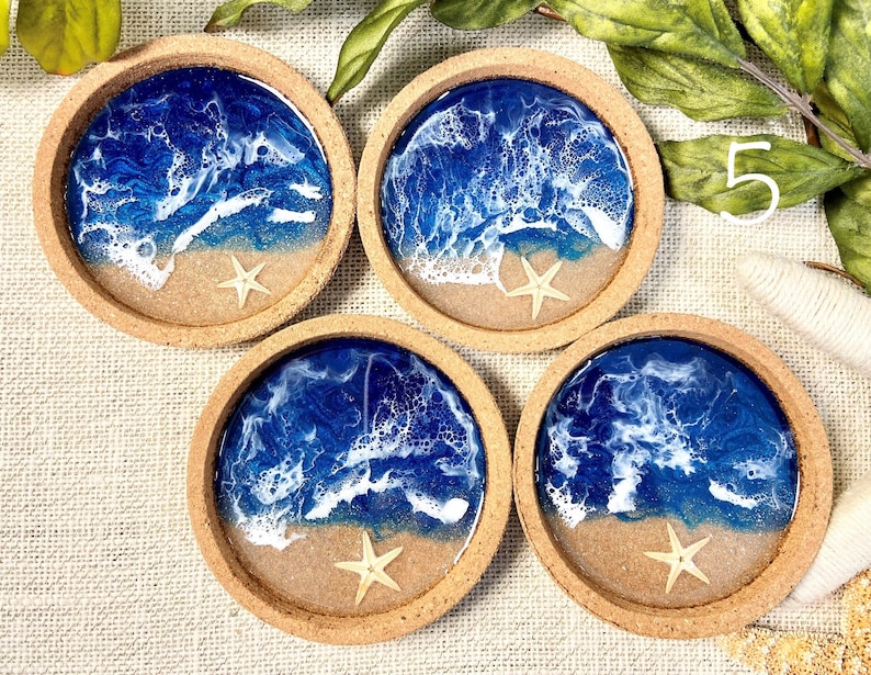 May include: Four circular coasters with a cork rim, each featuring a resin art design of a blue ocean with white sea foam waves crashing onto a sandy beach. A small, light-colored starfish is embedded in the sand section of each coaster.