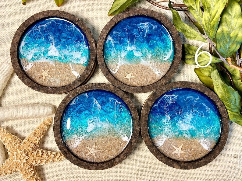 May include: Four circular coasters with dark brown cork frames. Each coaster features a resin ocean scene with a sandy beach, a small beige starfish, and swirling blue and turquoise water with white foam waves crashing against the shore.