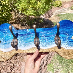 May include: A wooden dog bone shaped wall hook with three metal hooks. The bone is painted with blue and white resin to resemble an ocean wave.