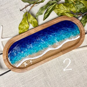 May include: A wooden serving tray with a blue and green resin ocean wave design. The tray has a metal handle and two small white seashells.