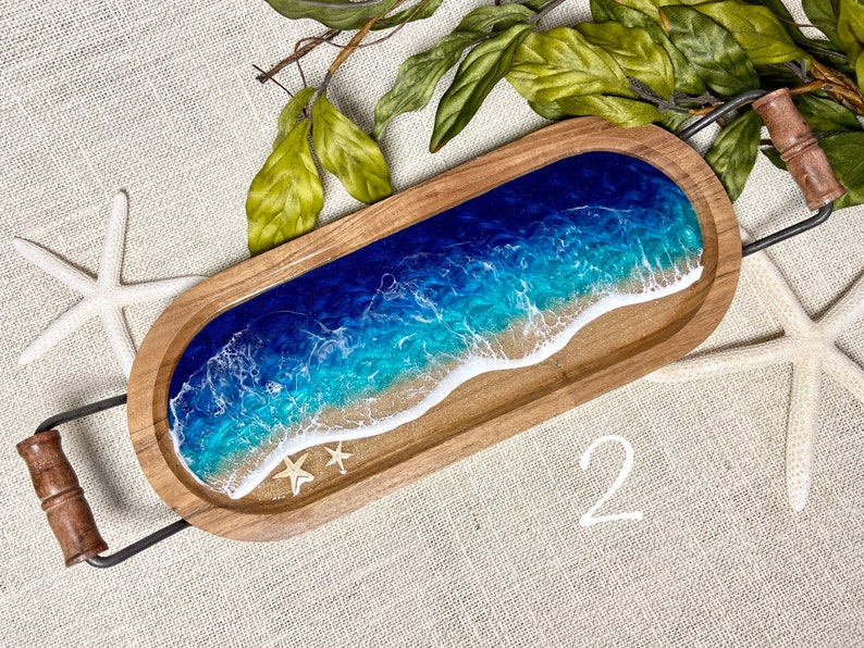 May include: A wooden serving tray with a blue and green resin ocean wave design. The tray has a metal handle and two small white seashells.