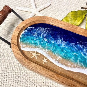 May include: A wooden serving tray with a resin ocean scene. The tray features blue and turquoise water with white foam waves and a sandy beach. Two white starfish are on the beach.