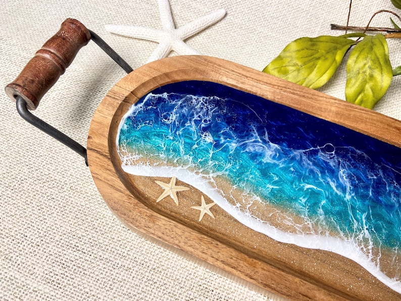 May include: A wooden serving tray with a resin ocean scene. The tray features blue and turquoise water with white foam waves and a sandy beach. Two white starfish are on the beach.
