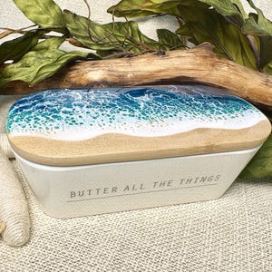 May include: A white butter dish with a wooden lid featuring a beach scene design. The lid has a sandy beach and ocean waves design. The words "BUTTER ALL THE THINGS" are printed on the front.