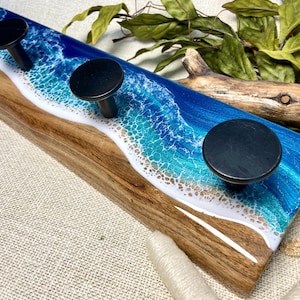 May include: Wooden wall hook with a blue and white resin ocean wave design. The hook has four black metal knobs.