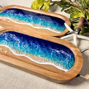May include: Two wooden serving trays with resin ocean wave designs. The trays are oval shaped and have metal handles. The resin design features blue, white, and light blue colors, resembling ocean waves crashing on a sandy beach. The trays are decorated with small white seashells.