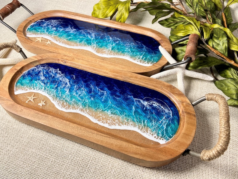 May include: Two wooden serving trays with resin ocean wave designs. The trays are oval shaped and have metal handles. The resin design features blue, white, and light blue colors, resembling ocean waves crashing on a sandy beach. The trays are decorated with small white seashells.