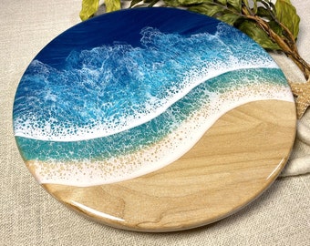 May include: Round wooden serving board with an ocean wave design. The top features a deep blue ocean, transitioning to turquoise and white waves, and a sandy beach base. The board is approximately 12 inches in diameter.