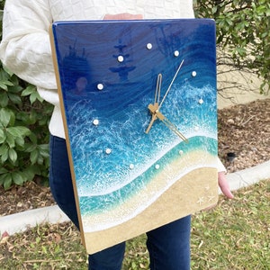 May include: A square resin clock with a beach scene. The clock face is blue with white waves and a sandy beach. The clock hands are gold.