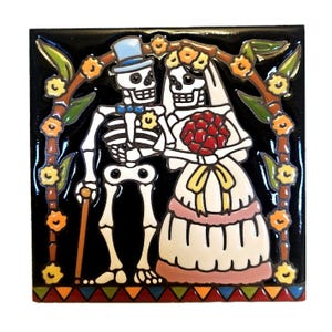 May include: Square ceramic tile featuring a Day of the Dead wedding scene. The bride and groom are skeletons under a floral arch. The bride holds a red bouquet, and the groom wears a top hat and carries a cane. The tile has a black background.