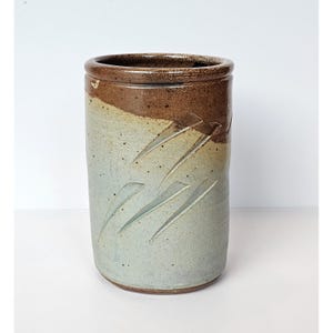 May include: A cylindrical ceramic vessel featuring a two-tone glaze with a dark brown rim and a light blue-grey body. The surface is decorated with carved diagonal slash marks that reveal the underlying clay texture across the exterior.