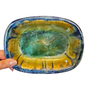 May include: An oval-shaped decorative dish with a unique, colorful design. The dish features a blend of blue, yellow, and green hues, creating a striking visual effect. The interior has a textured, crystalline appearance, adding depth and interest to the piece.