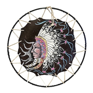May include: A Native American headdress painted on a brown leather circle with a black wooden frame. The headdress is white with black spots and pink feathers. The face is painted in shades of brown, pink, and white. The text "RTBP '13" is visible in the lower right corner.