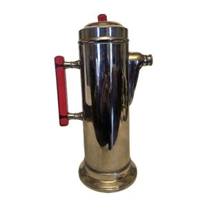 May include: A vintage chrome coffee percolator with a red handle and top accent. The cylindrical body has a spout and a base. The handle is a bright red color. The percolator is made of shiny metal.