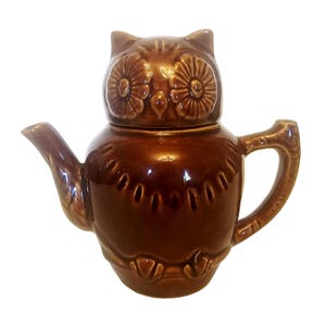 May include: A brown ceramic teapot shaped like an owl. The teapot has a lid that forms the owl's head, with detailed eyes and ear tufts. The body of the teapot has a rounded shape with a handle and spout.