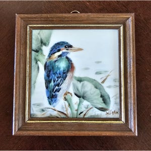 May include: A framed painting of a bird with blue, teal, and brown feathers, perched on a green plant. The artwork is set against a white background with a few water plants. The frame is made of brown wood.