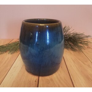 May include: A ceramic vase with a glossy, blue glaze that fades to a darker brown at the rim. The vase has a rounded, cylindrical shape. A sprig of greenery rests beside the vase on a wooden surface.