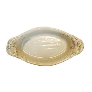 May include: Oval-shaped ceramic serving dish with a light cream color. The dish features raised relief details of seashells and a pinkish-tan seahorse on each end. The rim has a subtle, darker band.