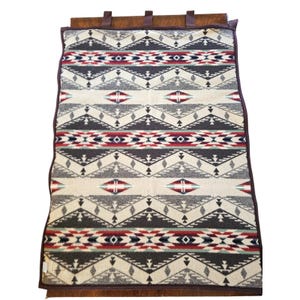 May include: A rectangular blanket with a geometric pattern in cream, gray, red, and navy blue. The blanket has a brown leather trim and straps. The pattern features diamond and arrow shapes. The blanket is folded over a wooden surface.