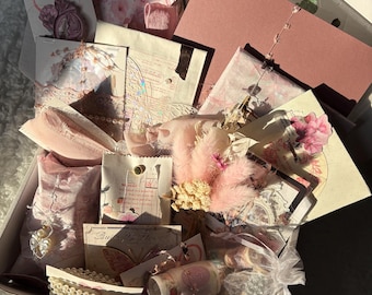 May include: A box filled with various craft supplies in shades of pink and white. The contents include decorative paper, dried flowers, ribbons, washi tape, and small embellishments. The box is open, revealing the contents arranged inside.