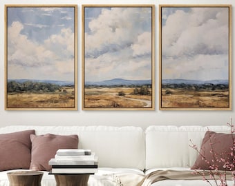 May include: Three framed landscape paintings with a muted color palette depicting a field with trees and a cloudy sky. The paintings are hung on a white wall above a white sofa with pillows and a throw blanket.