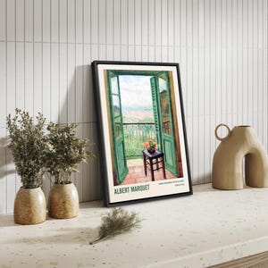 May include: A framed print of a painting by Albert Marquet titled "Window overlooking the Bay of Algiers". The painting depicts a view from a balcony with open green doors, a small table with a vase of flowers, and a view of the sea and coastline.