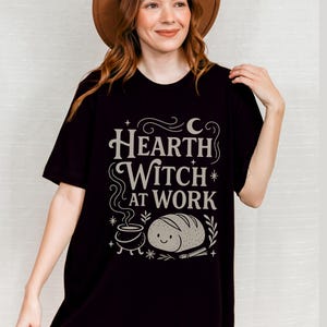 May include: Black t-shirt with the text "Hearth Witch at Work" in a whimsical font. The design includes a crescent moon, stars, a cauldron, and a smiling loaf of bread. The shirt is worn by a person wearing a brown hat.