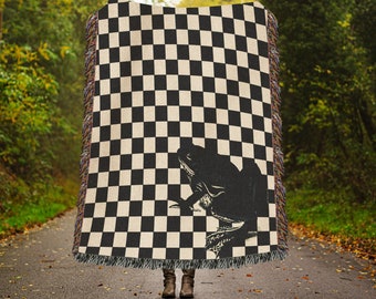 May include: A black and white checkered woven throw blanket with a black silhouette of a frog on one side. The blanket is draped over a person's shoulders.