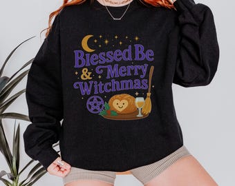 Może przedstawiać: Ciemnoszara bluza z napisem "Blessed Be & Merry Witchmas" w kolorze fioletowym i złotym. Projekt zawiera półksiężyc, gwiazdy, pentagram, bochenek chleba z sercem, kieliszek wina i miotłę.