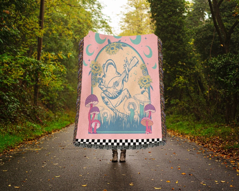 May include: A pink woven tapestry throw blanket with a whimsical design featuring a frog playing a guitar, surrounded by mushrooms and sunflowers. The design is set against a blue and green background with a crescent moon.