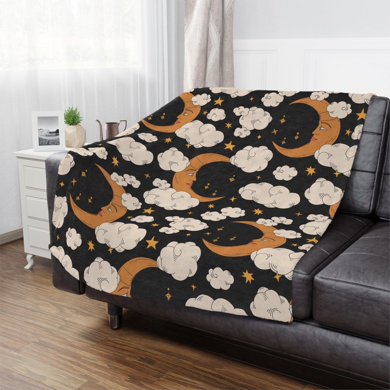 May include: A black throw blanket with a repeating pattern of orange crescent moons with faces, white clouds, and gold stars. The blanket is draped over a black couch.
