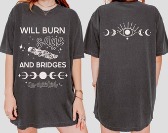 May include: Black oversized t-shirt with white text that reads "Will burn sage and bridges as needed". The back of the shirt has a white eye with rays and three crescent moons.