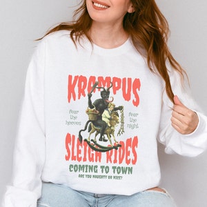 May include: White sweatshirt with a red graphic print of Krampus riding a sleigh with a child. The text reads "Krampus Sleigh Rides Coming to Town Are You Naughty or Nice?"