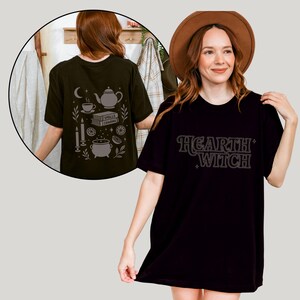 Może przedstawiać: Czarny t-shirt z napisem "Hearth Witch" w stylu retro. Koszulka ma luźny krój. Tył koszulki, widoczny w okrągłej wstawce, przedstawia wzór z czajnikiem, filiżanką i innymi mistycznymi elementami.