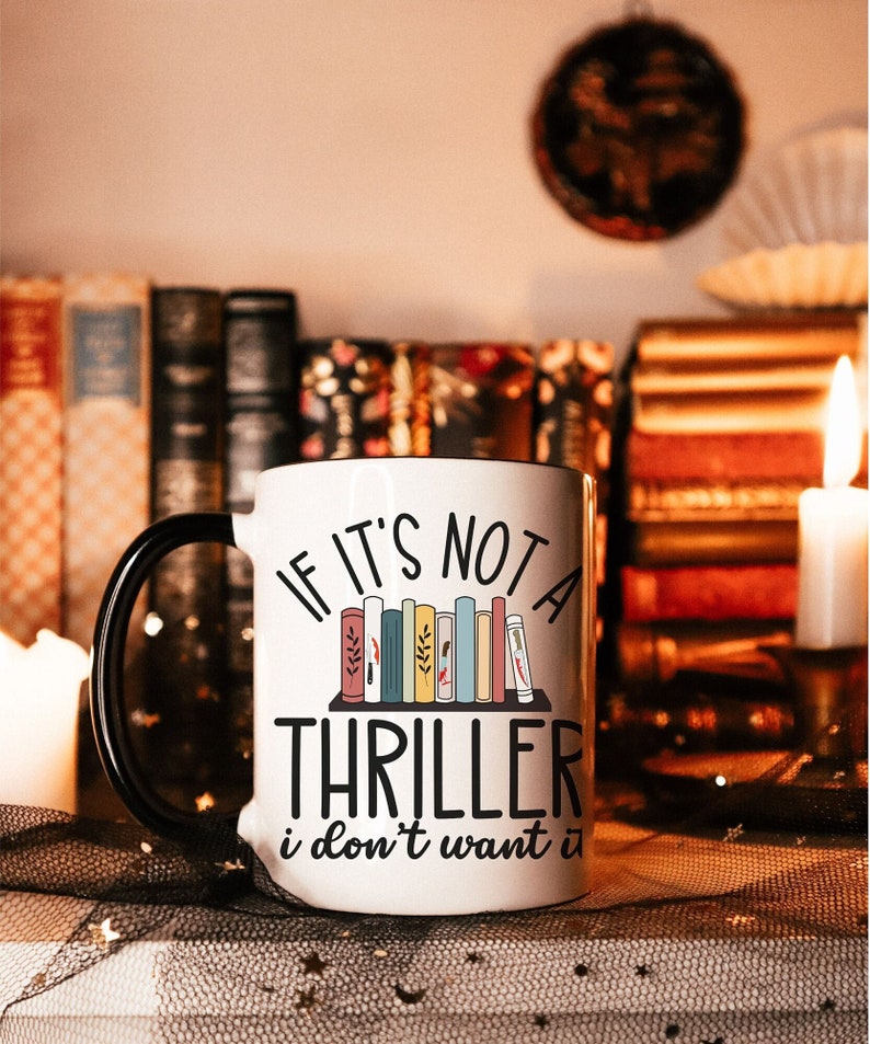 A white mug with a black-colored handle with a quote that says "If it's not a thriller I don't want it" with a shelf filled with books. There's a lit candle with books behind the mug.