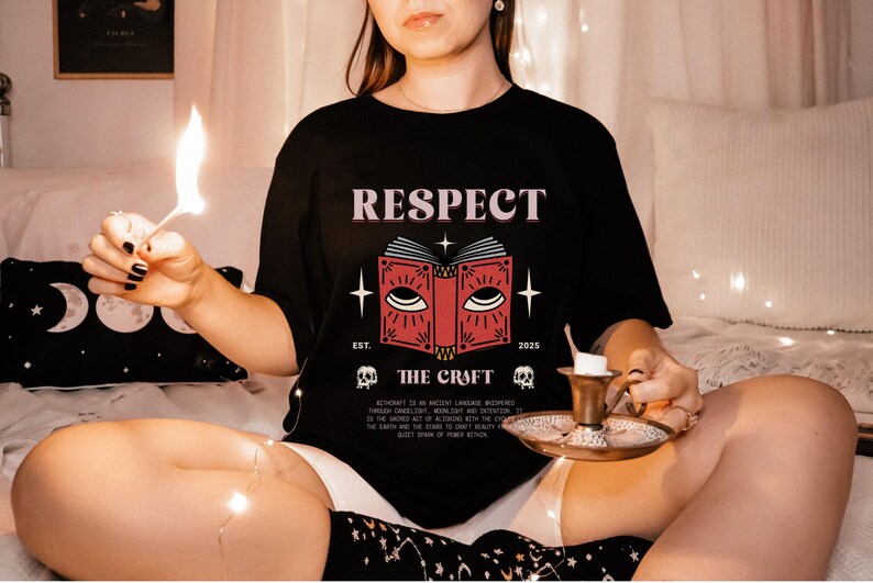 May include: Black t-shirt with the word "RESPECT" above a red book graphic with eyes, stars, and the text "THE CRAFT". The shirt also features the year "2025" and skull graphics. A lit match and candle holder are also visible.