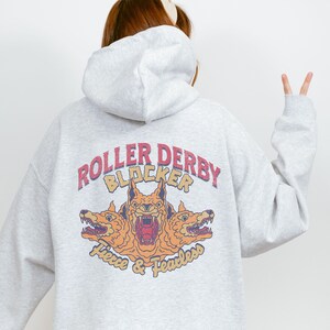 Può includere: Felpa con cappuccio grigia con una stampa grafica di tre teste di lupo con il testo "Roller Derby Blocker Fierce & Fearless" sul retro.