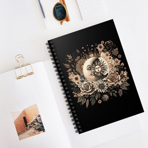 May include: Black spiral-bound notebook featuring a celestial design. The cover showcases a detailed illustration of a crescent moon and a sun, surrounded by floral elements in shades of beige and brown. The notebook is open on a white surface.