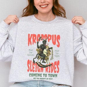 May include: Gray sweatshirt with a red graphic print of a Krampus figure riding a rocking horse with a child. The text reads "Krampus Sleigh Rides Coming to Town Are You Naughty or Nice?"