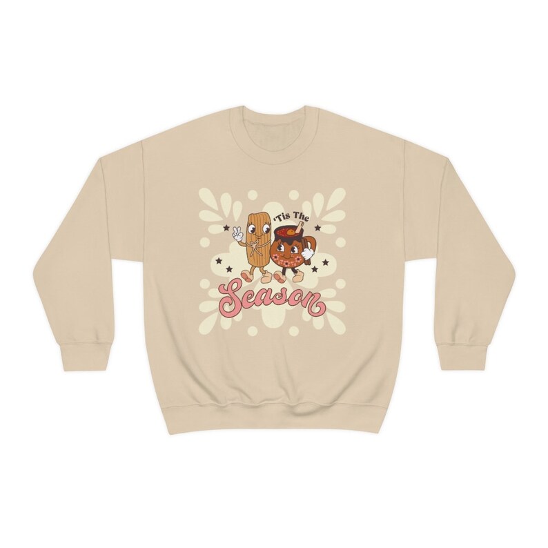 Op de afbeelding: Een beige sweatshirt met een cartoon afbeelding van een kaneelstokje en een mok warme chocolademelk. De tekst "Tis the Season" is in roze onder de cartoonfiguren gedrukt.