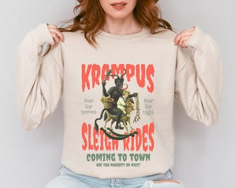 May include: A beige sweatshirt with a graphic print of a red-skinned demon riding a rocking horse. The text on the graphic reads "Krampus Sleigh Rides Coming to Town Are You Naughty or Nice?"