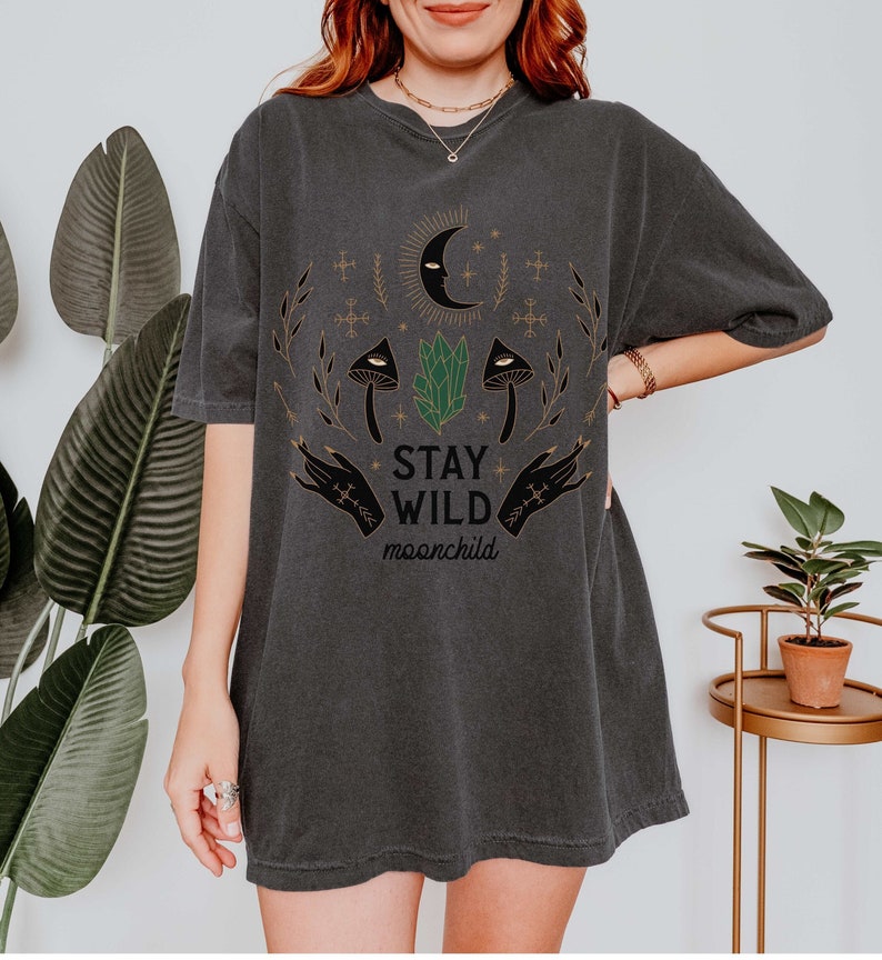 K&ouml;nnte beinhalten: Ein dunkelgraues Oversize-T-Shirt mit einem grafischen Druck eines zunehmenden Mondes, zwei Pilzen, einem Kristall und zwei H&auml;nden mit dem Text "STAY WILD moonchild".