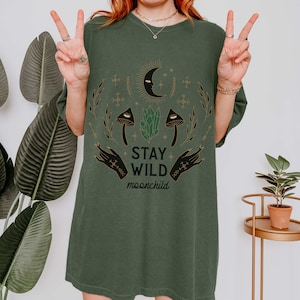 K&ouml;nnte beinhalten: &Uuml;bergr&ouml;&szlig;en-T-Shirt in Gr&uuml;n mit einem grafischen Druck eines Halbmondes, Sternen, Kristallen, Pilzen und H&auml;nden. Der Text "Stay Wild moonchild" ist auf das Shirt gedruckt.