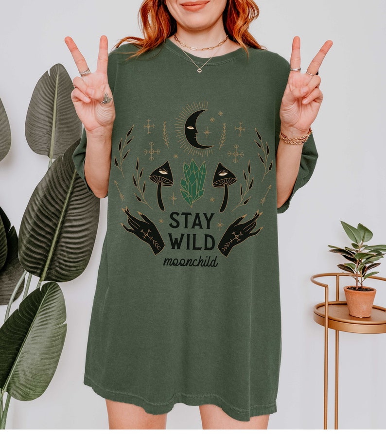 K&ouml;nnte beinhalten: &Uuml;bergr&ouml;&szlig;en-T-Shirt in Gr&uuml;n mit einem grafischen Druck eines Halbmondes, Sternen, Kristallen, Pilzen und H&auml;nden. Der Text "Stay Wild moonchild" ist auf das Shirt gedruckt.