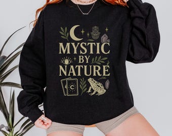 May include: A black sweatshirt with the text "MYSTIC BY NATURE" in a stylized font. The design includes a crescent moon, eye, hand, crystals, tarot cards, and a frog. The sweatshirt is a dark charcoal color.