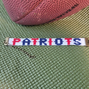 May include: A beaded bracelet featuring a white background with the word PATRIOTS spelled out in alternating red and blue beads. The bracelet includes bronze-toned metal end caps and a matching metal chain extender for adjustable sizing.
