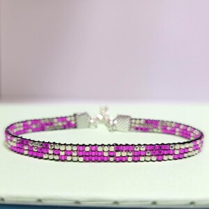 May include: A beaded bracelet with a repeating pattern of magenta and silver beads. The bracelet has a silver clasp and is displayed on a light green surface. The beads are small and evenly spaced, creating a textured look.