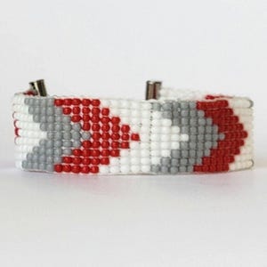 Könnte beinhalten: Ein Armband aus Perlen mit einem geometrischen Muster. Das Armband hat ein sich wiederholendes Design aus roten, grauen und weißen Perlen. Das Armband hat silberfarbene Verschlüsse an jedem Ende. Das Armband ist ein Modeaccessoire.