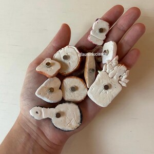 Pu&ograve; includere: Una collezione di calamite fatte a mano in argilla di varie forme, tra cui cerchi, rettangoli e forme organiche. Le calamite sono principalmente bianche con tocchi di marrone, arancione e nero. Ogni calamita ha un piccolo magnete color argento sul retro.