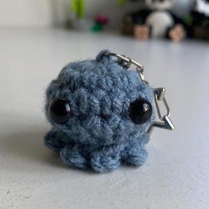 May include: A small, crocheted octopus keychain in a muted blue color. The octopus has two large, black button eyes and a silver-colored metal keychain attachment. The tentacles are slightly visible, and the overall design is soft and rounded.
